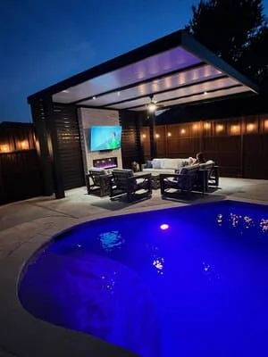 Lit pool with blue LED lights, modern pergola with shade panels, patio seating, and landscape lighting at dusk