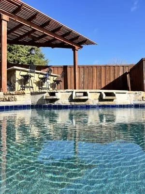 Pool deck with pergola, concrete surround, and wood privacy fence under clear sky