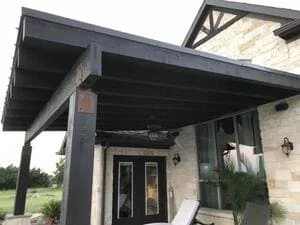 Steel pergola with black posts and beams over patio, attached to stone house